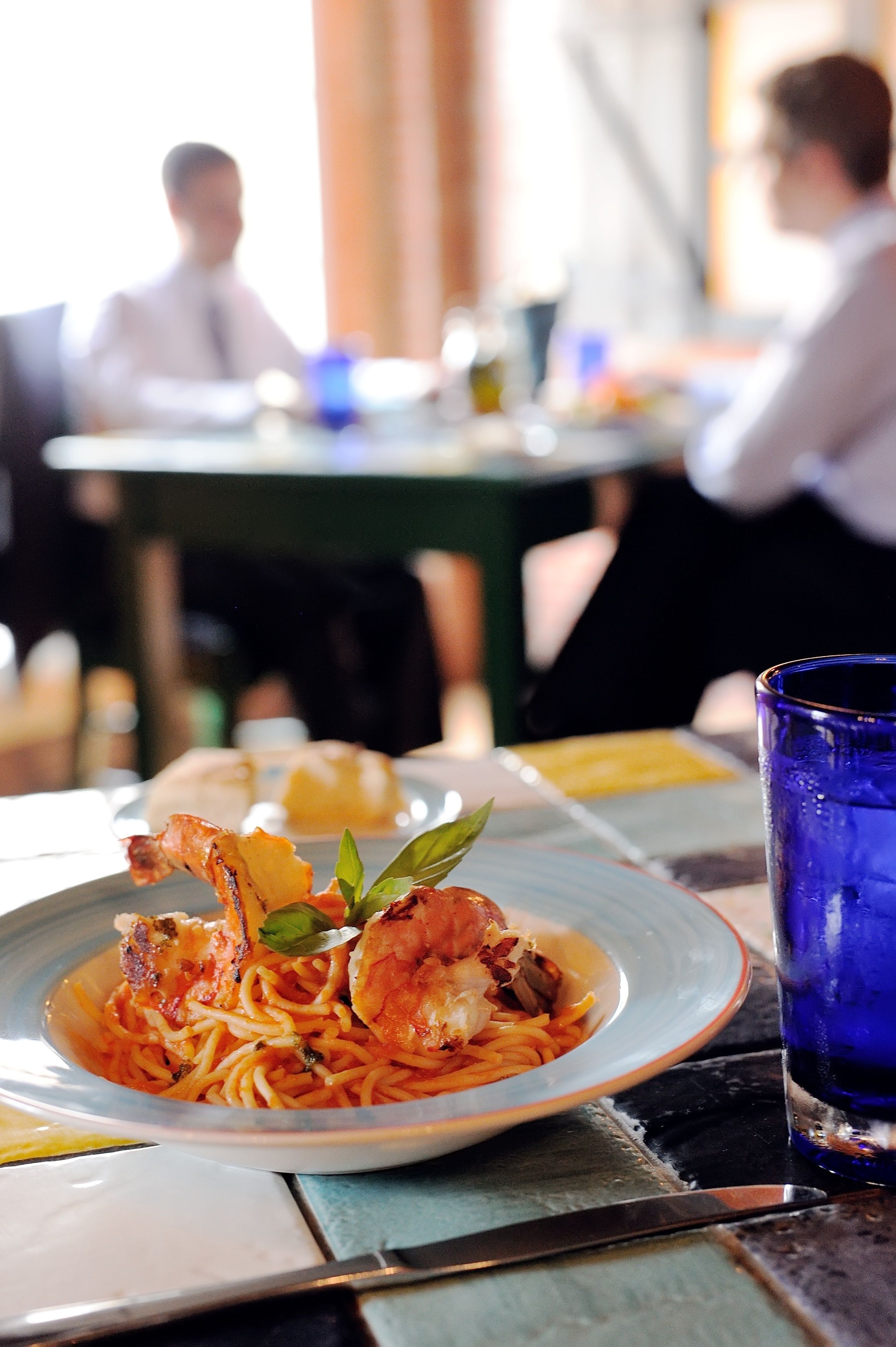 Uma mesa de jantar com um prato de massa e camarão, um copo azul e uma faca.