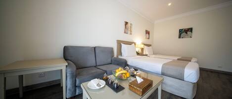 A hotel room with a sofa, a bed, a glass coffee table with fruits and a box, and a small table with a lamp.