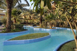 A pool area with multiple water features surrounded by palm trees and a wooden bridge. - Montrose A Mayfair Collection Hotel (Kuwait City)