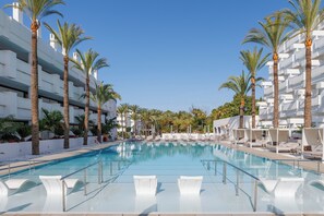 Una piscina grande con sillones blancos y palmeras, rodeada de edificios blancos.