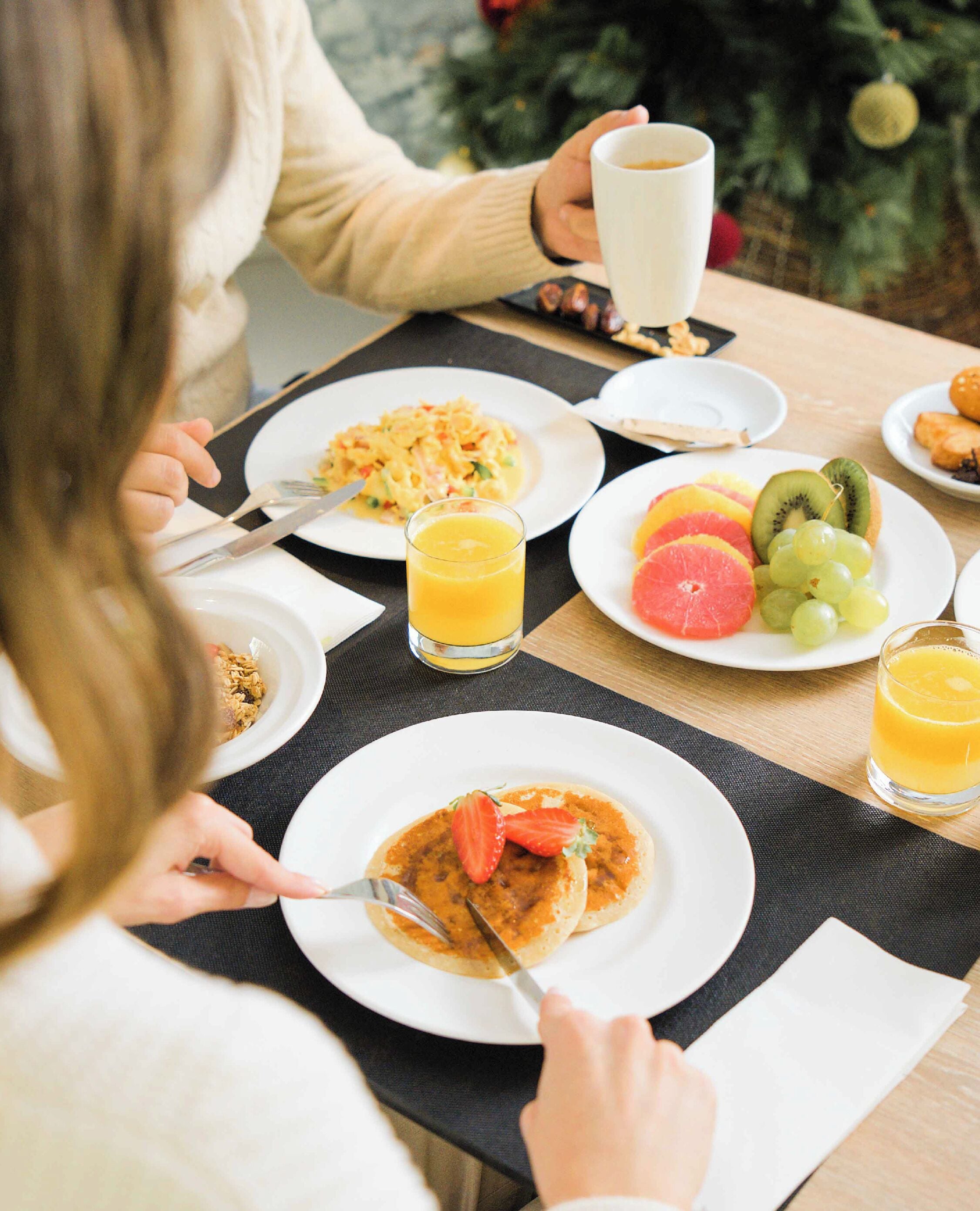 Une table dressée pour le petit-déjeuner, avec des assiettes remplies de nourriture, un verre de jus d’orange et un sapin de Noël en arrière-plan.