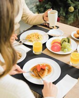 Une table dressée pour le petit-déjeuner, avec des assiettes remplies de nourriture, un verre de jus d’orange et un sapin de Noël en arrière-plan.