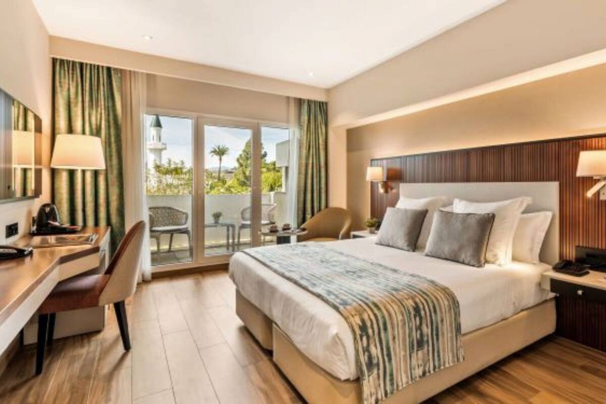 Ein modernes Hotelzimmer mit einem großen Bett, einem Schreibtisch und einem Sessel. Es gibt einen Balkon mit Blick ins Freie.