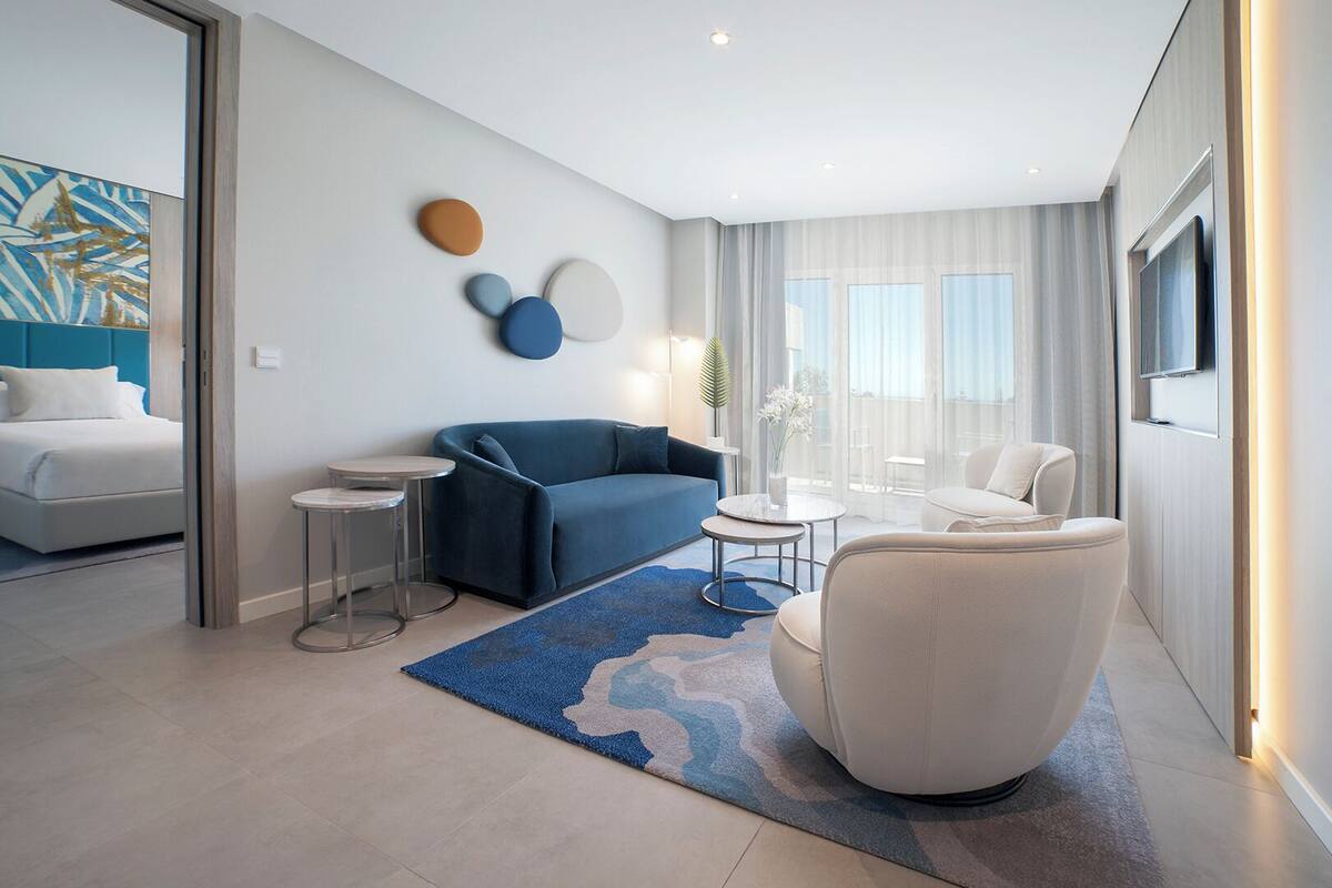 Ein modernes Hotelzimmer mit einem Bett, einem blauen Sofa, einem weißen Sessel, einem kleinen runden Tisch und einem großen Fenster mit Vorhängen.