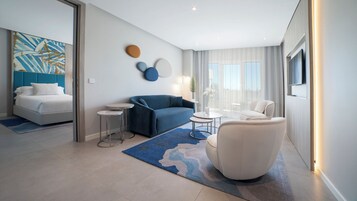 Een moderne hotelkamer met een bed, een blauwe bank, een witte fauteuil, een kleine ronde tafel en een groot raam met gordijnen.