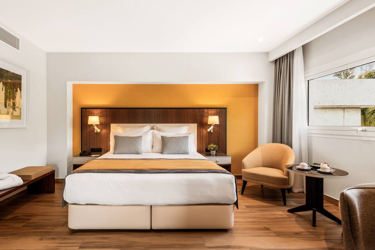 Ein modernes Hotelzimmer mit einem großen Bett, Nachttischen, einem Sessel, einem kleinen Tisch mit Tassen und einem Fenster mit Blick.