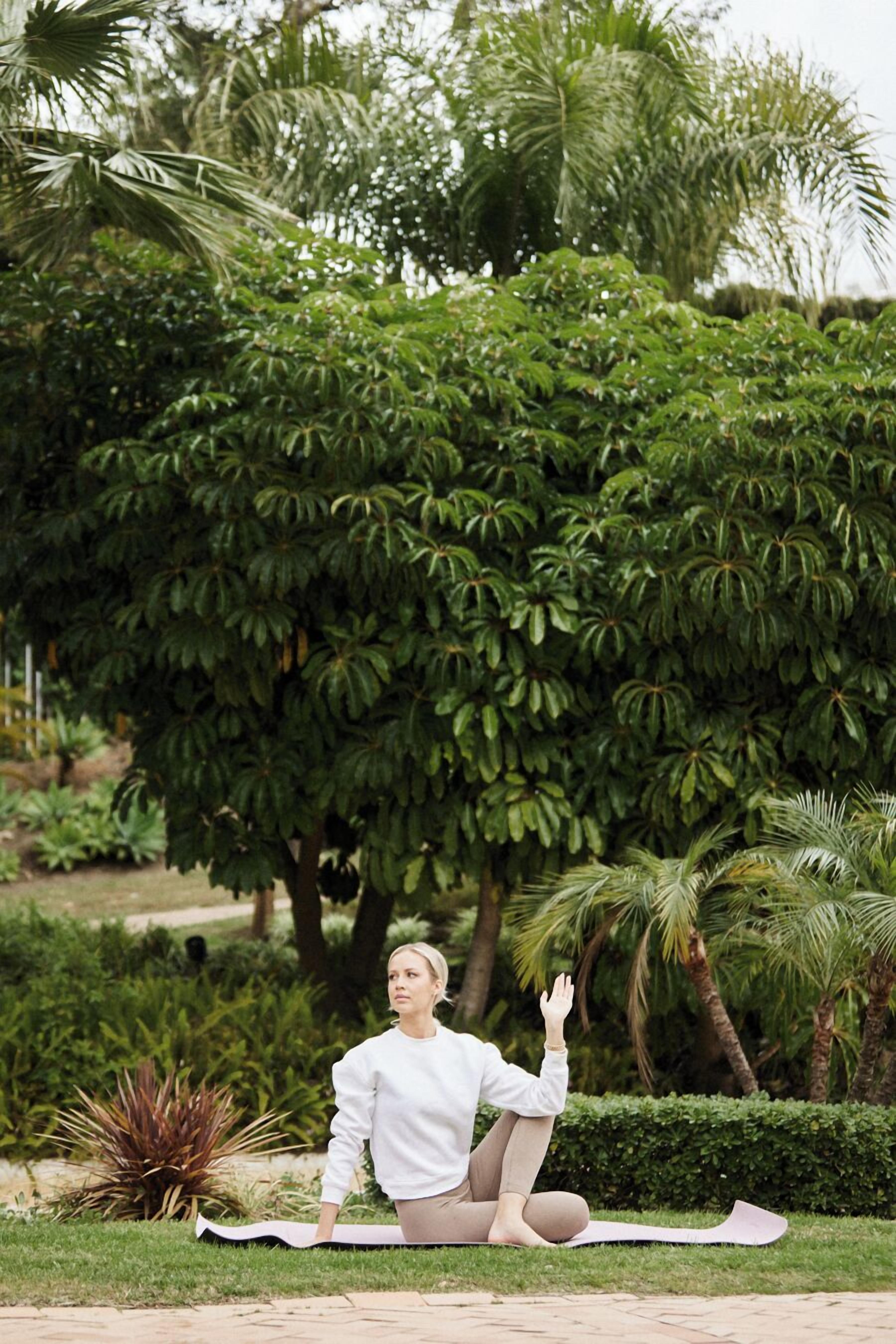 A person sitting on a mat outdoors with trees and a hedge in the background.