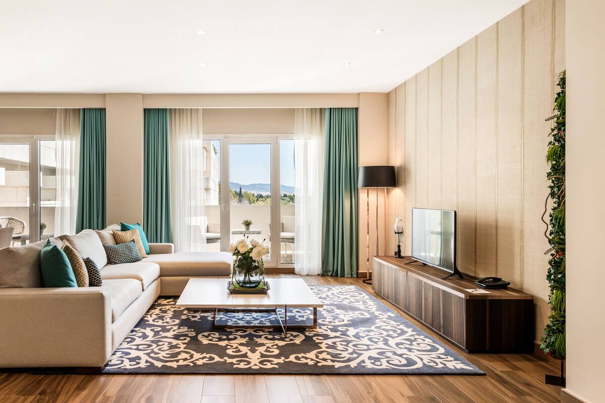 Ein modernes Wohnzimmer mit Sofa, Couchtisch und Fernseher. Es gibt einen Balkon mit Vorhängen und Blick ins Freie.