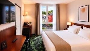 A hotel room with a bed, a desk, a chair, a TV, a balcony with a view of buildings, and a door. - Mercure Lourdes Imperial (Lourdes)