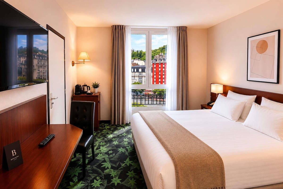 A hotel room with a bed, a desk, a chair, a TV, a balcony with a view of buildings, and a door.