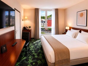 A hotel room with a bed, a desk, a chair, a TV, a balcony with a view of buildings, and a door. - Mercure Lourdes Imperial (Lourdes)