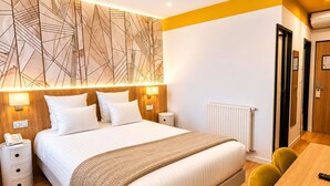 Una habitación de hotel moderna con una cama grande, lámparas de noche, un cabecero de madera, una mesita de noche con un teléfono, un radiador y un escritorio de madera con una silla.