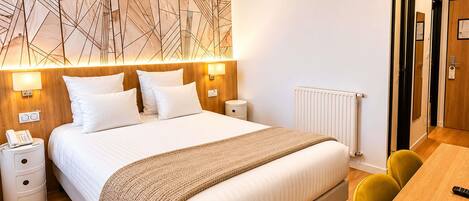 Una habitación de hotel moderna con una cama grande, lámparas de noche, un cabecero de madera, una mesita de noche con un teléfono, un radiador y un escritorio de madera con una silla.