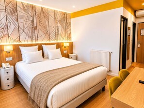 A modern hotel room with a large bed, bedside lamps, a wooden headboard, a bedside table with a phone, a radiator, and a wooden desk with a chair. - Mercure Lourdes Imperial (Lourdes)