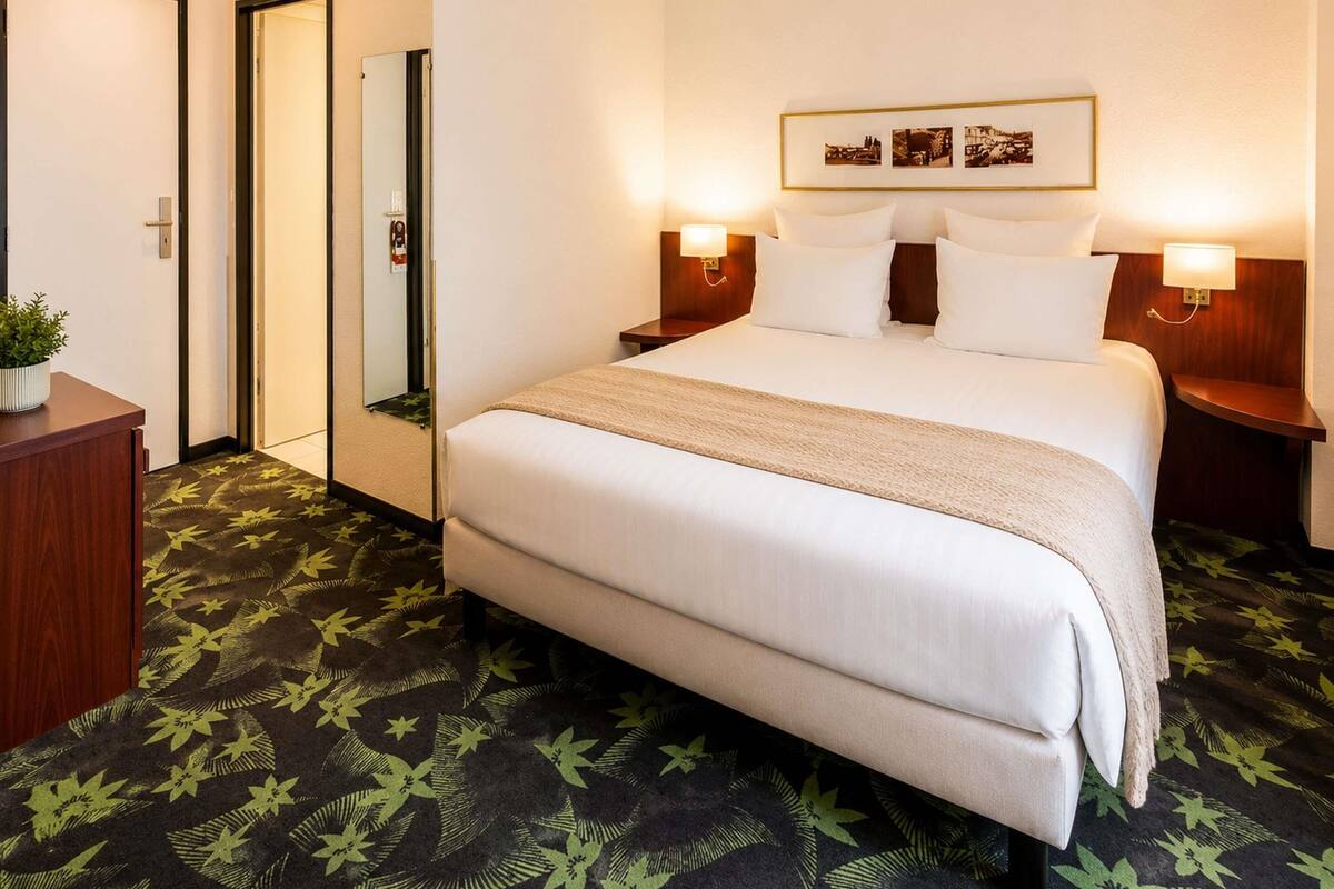 A hotel room with a bed, nightstands, a mirror, a framed picture on the wall, and a patterned carpet.