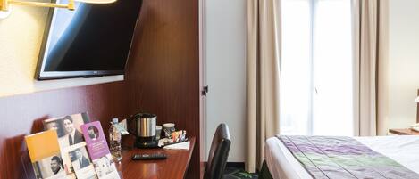 A hotel room with a bed, a desk with a lamp, a television, and a window with curtains.