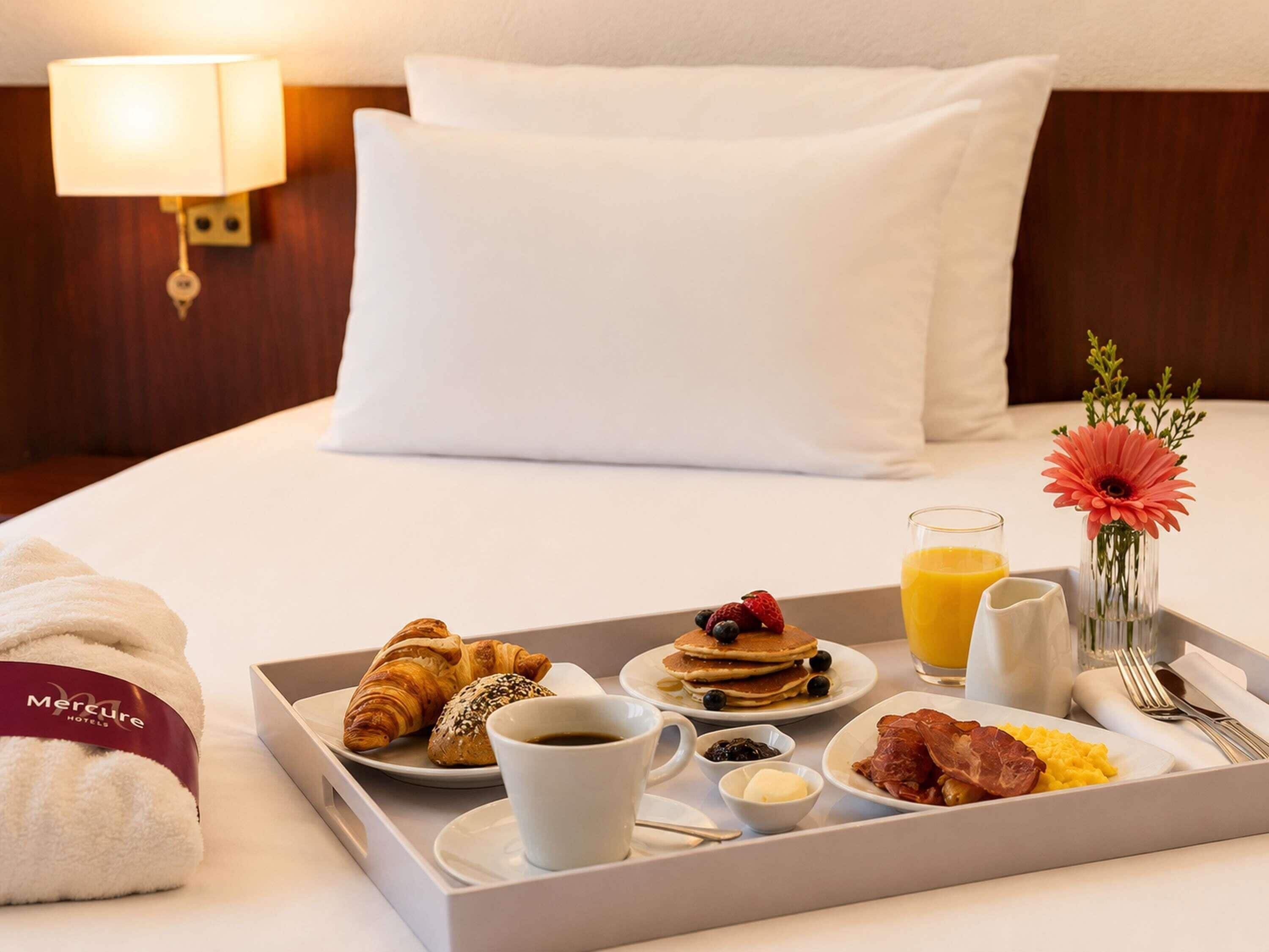 A hotel room with a neatly made breakfast tray, including croissants, pancakes, berries, scrambled eggs, bacon, toast, coffee, and orange juice.