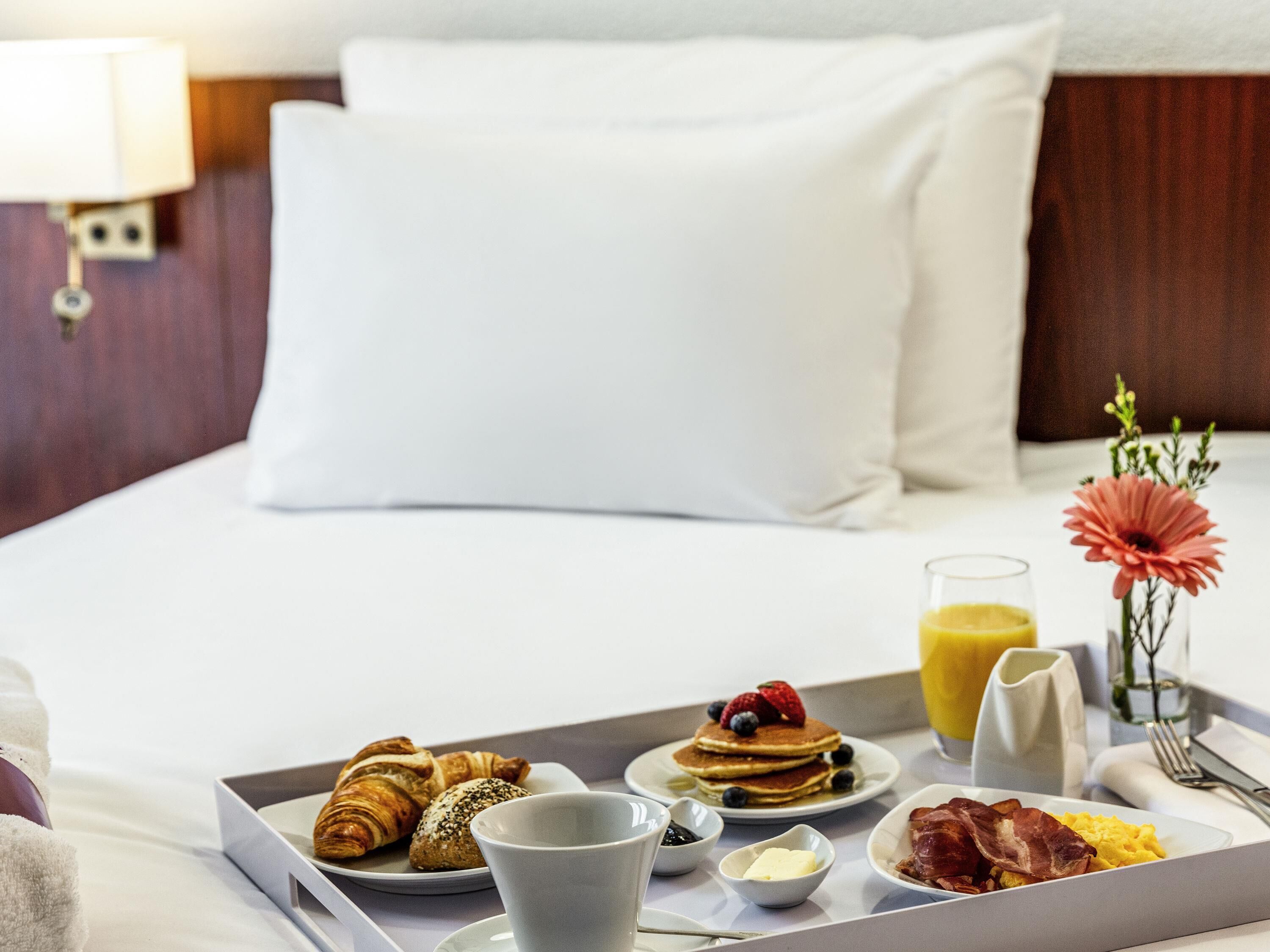 Un plateau de petit-déjeuner joliment préparé, avec des croissants, des crêpes, des fraises, des myrtilles et du bacon, posé sur un lit recouvert de draps blancs et doté d’une tête de lit en bois.