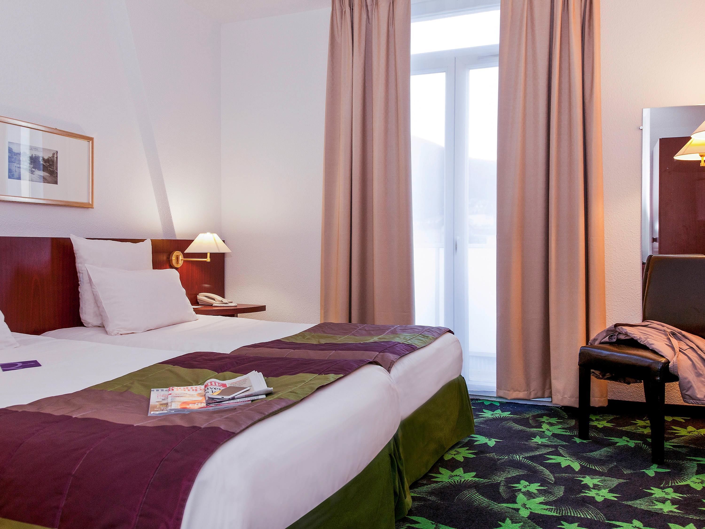 A hotel room with a large bed, a bedside table with a lamp, a chair, and a window with curtains.