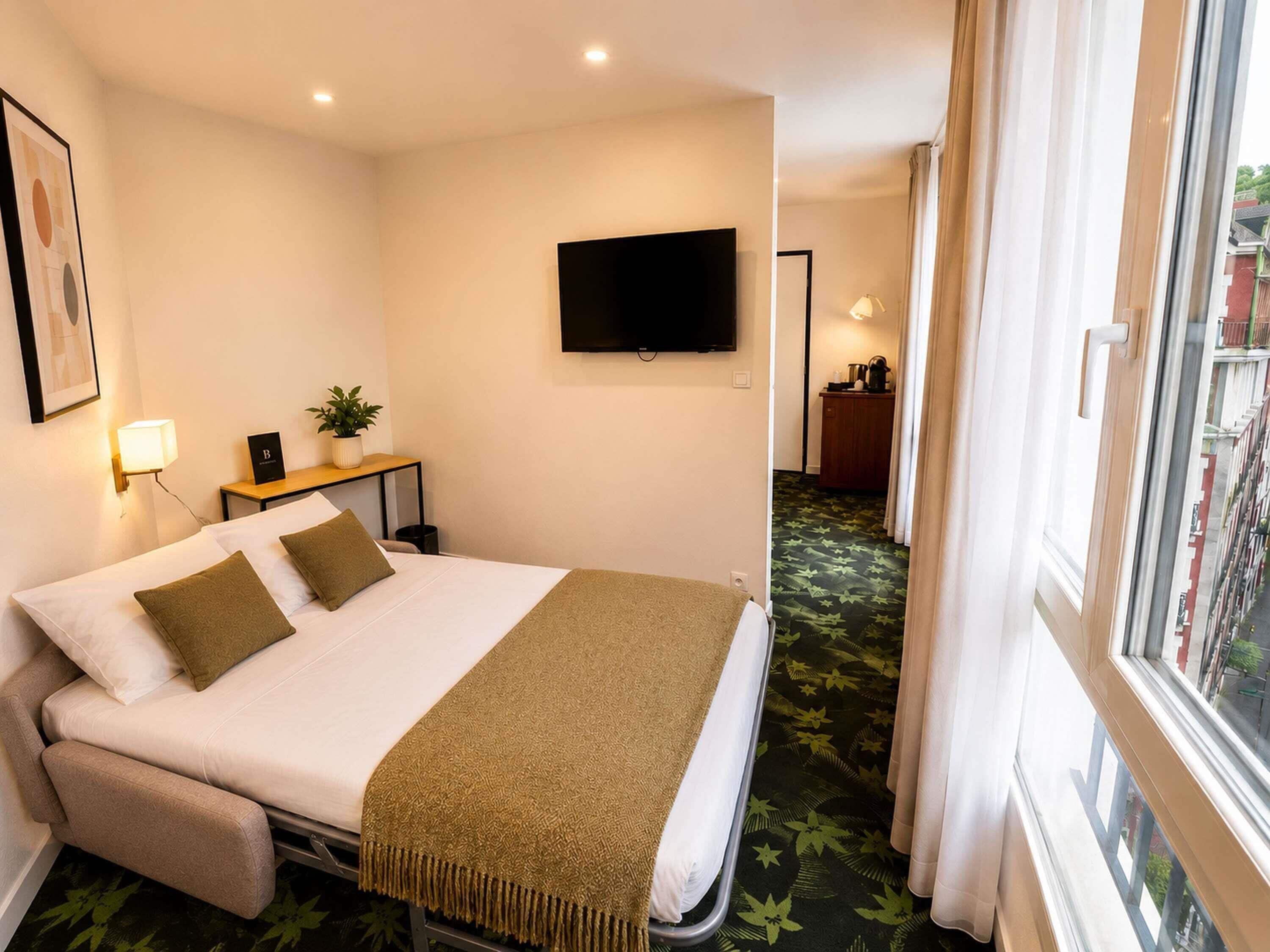 A hotel room with a bed, a TV mounted on the wall, a bedside table with a lamp, and a window with curtains.