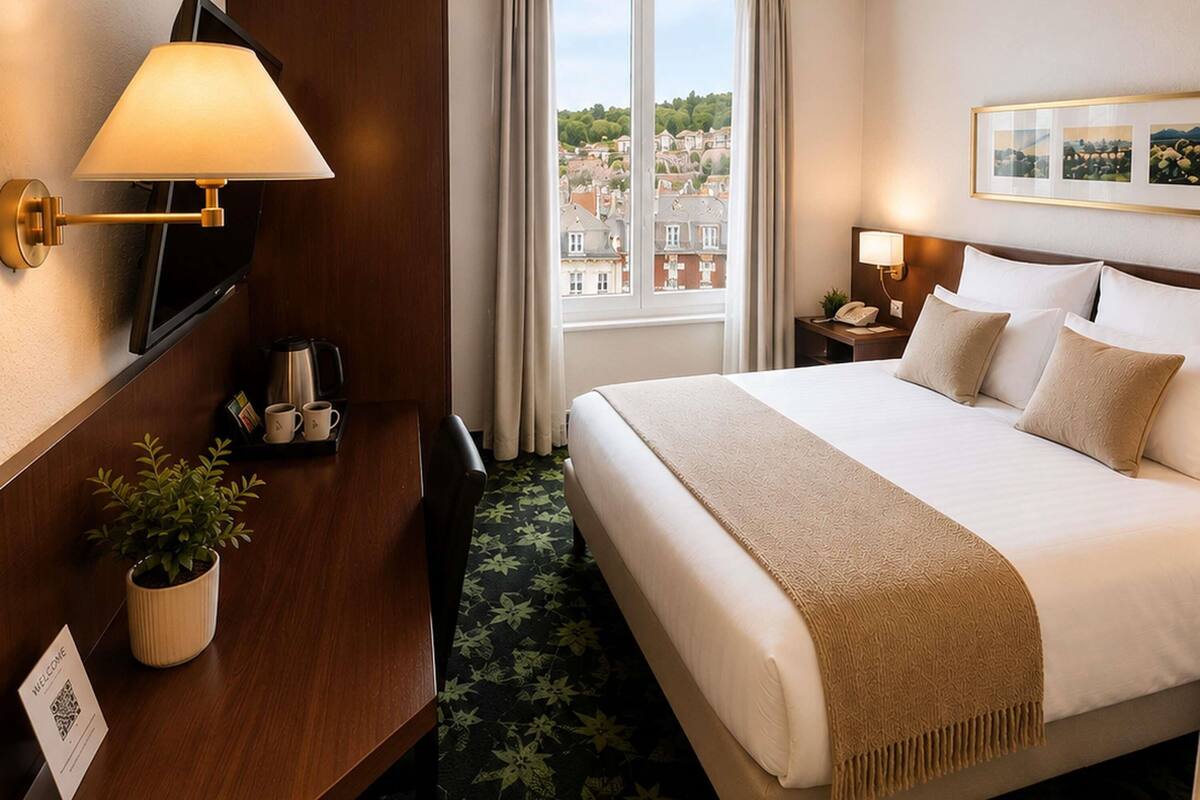 Habitación de hotel con cama, escritorio con cafetera, televisor, ventana con vista a la ciudad y un cuadro en la pared.