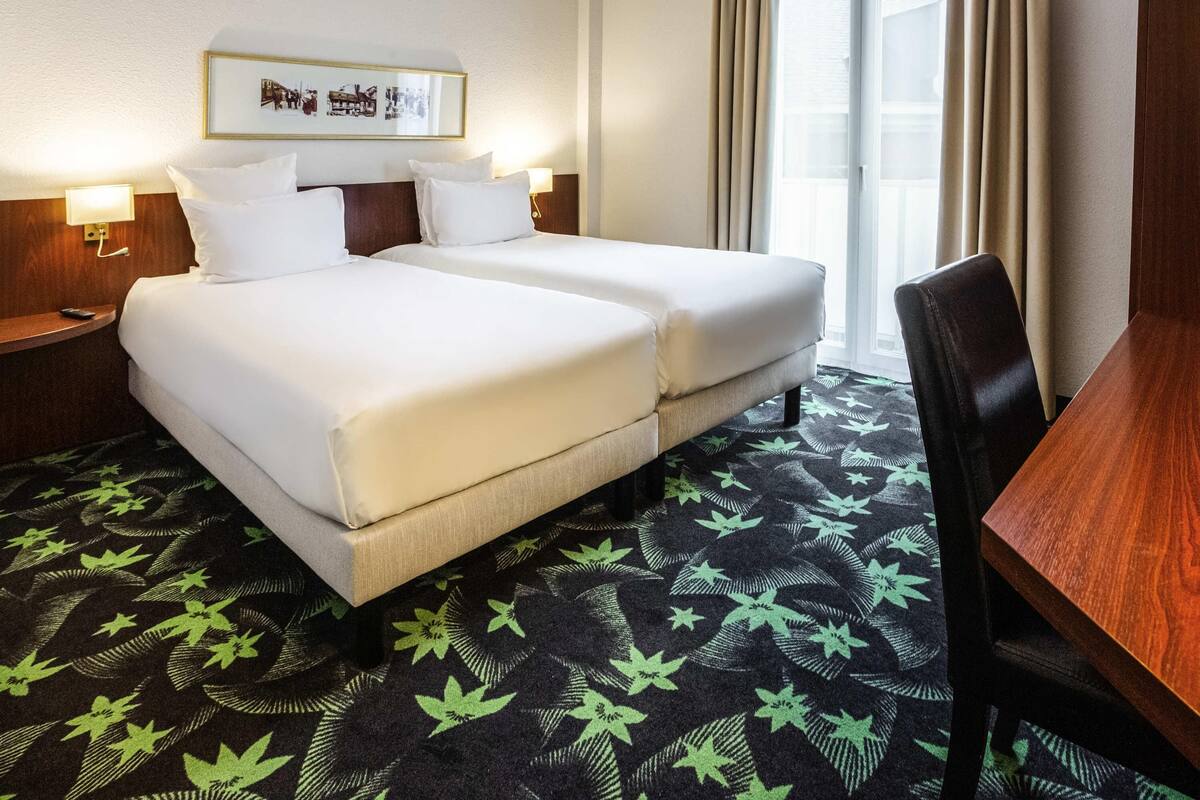 A hotel room with two beds, a desk, a chair, a framed picture on the wall, and a window with curtains.