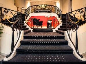 A grand staircase with ornate black railings and a red wall at the top. - Mercure Lourdes Imperial (Lourdes)