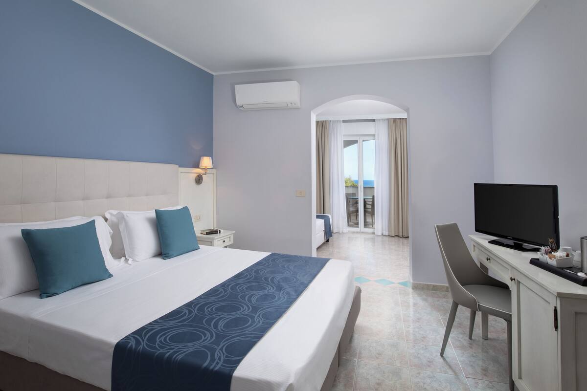 A hotel room with a large bed, a desk with a TV, and a view of the balcony.