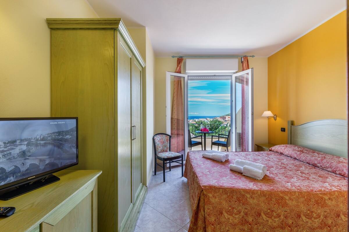 A hotel room with a bed, a television, a chair, a table, and a balcony with a view.