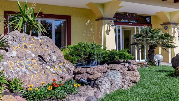Exterior de un hotel con jardín de rocas, fuente de agua y un letrero que dice 'Hotel Rosa del Viento'.
