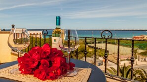 Una mesa con dos copas de vino y una botella, flores rojas y vista a la playa y el océano.