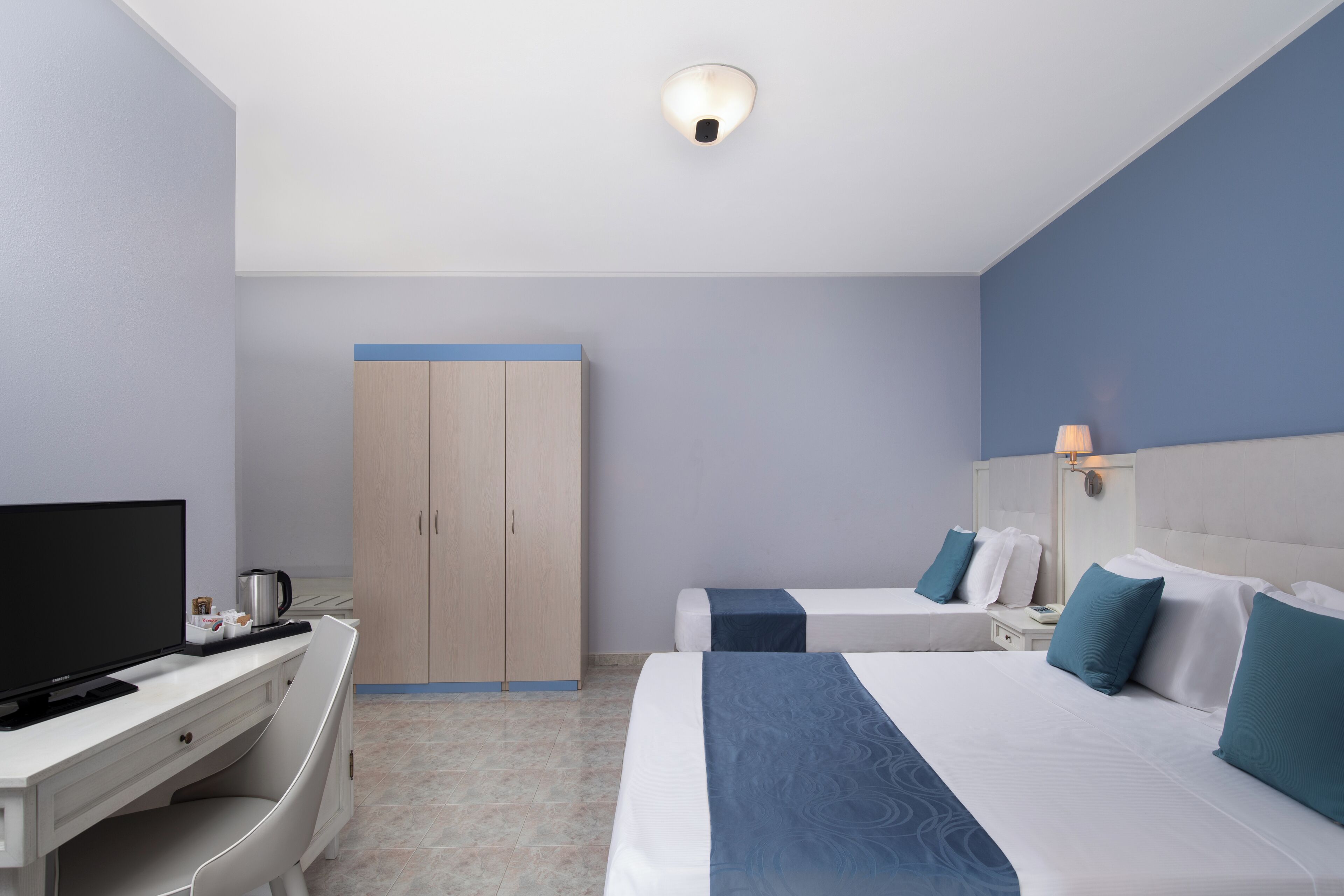 A hotel room with two beds, a desk with a TV, a wardrobe, and a ceiling light.