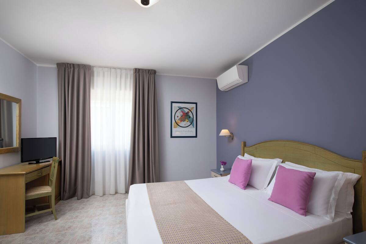 A hotel room with a bed, a desk, a chair, a TV, a wall-mounted air conditioner, and a framed picture.
