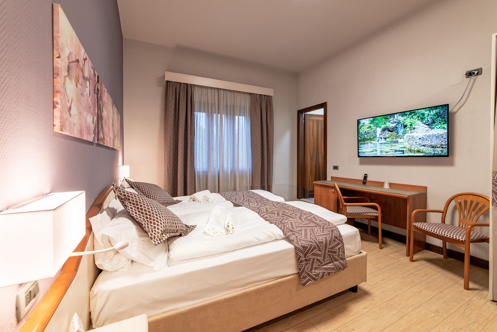 A hotel room with a bed, desk, chair, TV, and artwork on the wall.