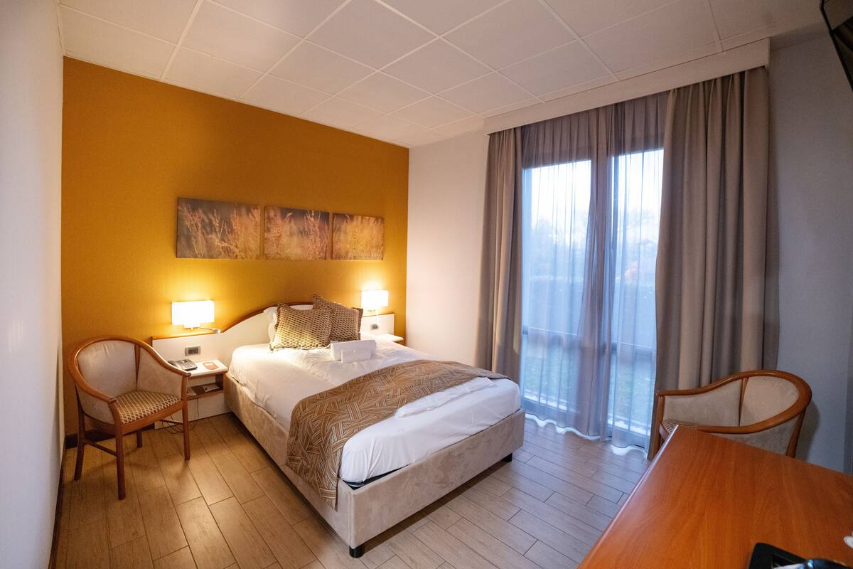 Una habitación de hotel con una cama, dos mesitas de noche con lámparas, una silla de madera, una mesa de madera y un amplio ventanal con cortinas.