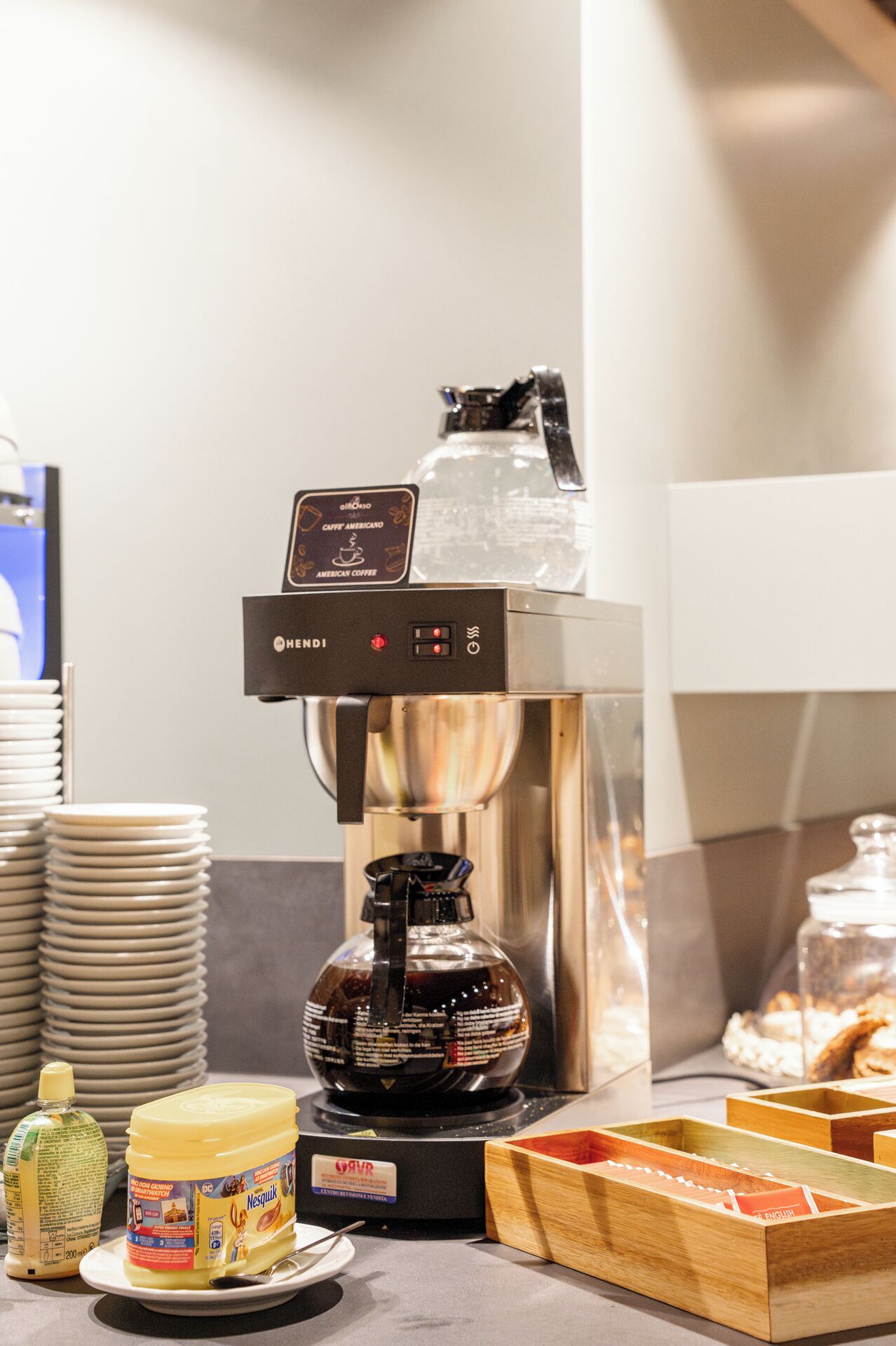 Eine Kaffeemaschine mit dem Hinweis für amerikanischen Kaffee, ein Stapel Teller, ein Nutella-Glas und eine Holzkiste mit Fächern.