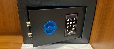 A black safe with a blue lock and keypad, placed on a wooden surface. Below it, a white paper labeled "LAVANDERIA LAUNDRY BAG" lists laundry instructions in multiple languages.