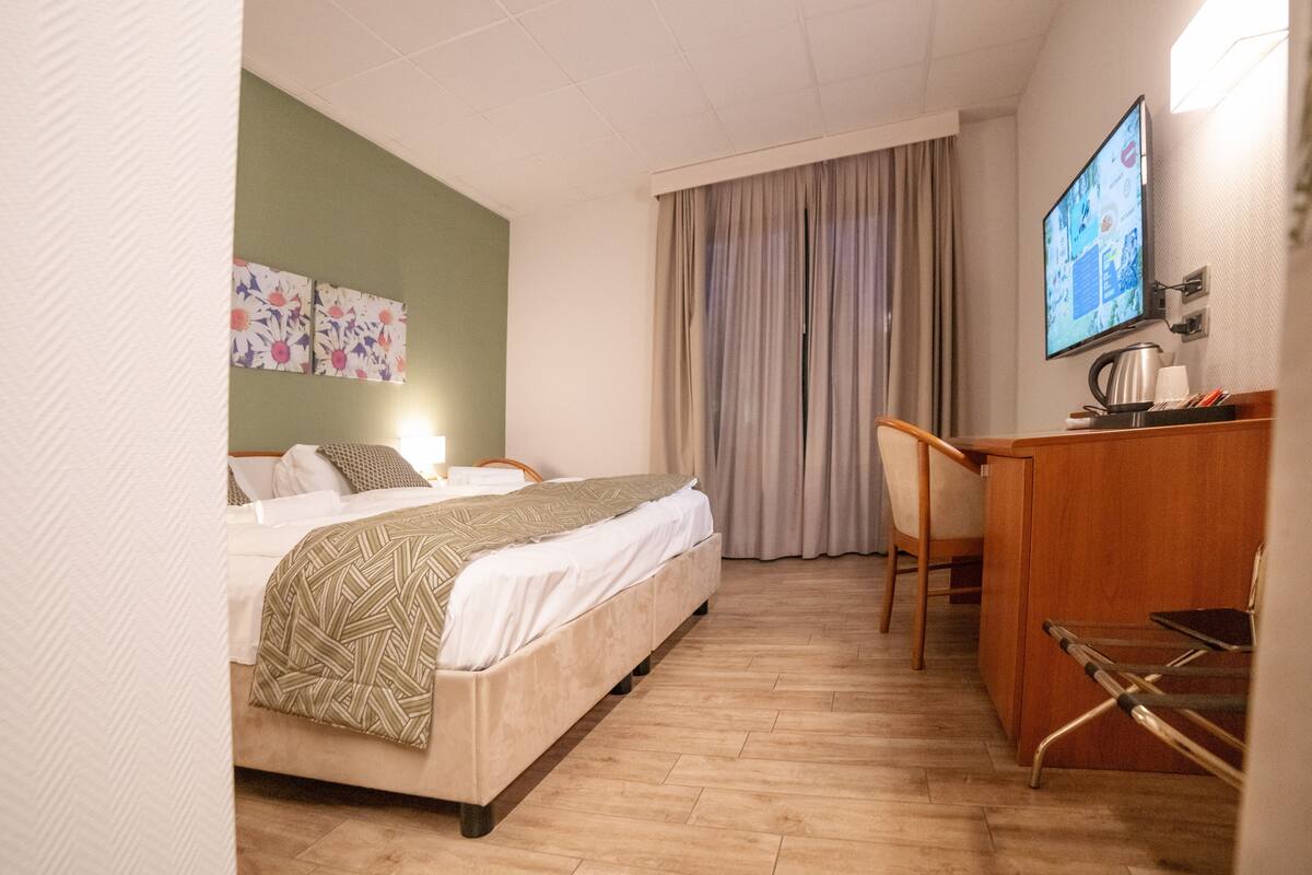Habitación de hotel con cama, escritorio, silla, televisor y ventana con cortinas.