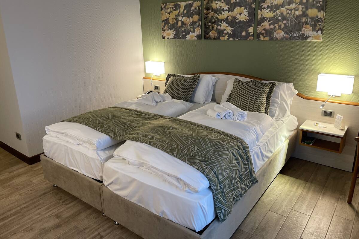 Una cama doble con una colcha estampada, dos lámparas de noche, una mesita de noche con un teléfono, y una obra de arte colgada en la pared sobre la cama.
