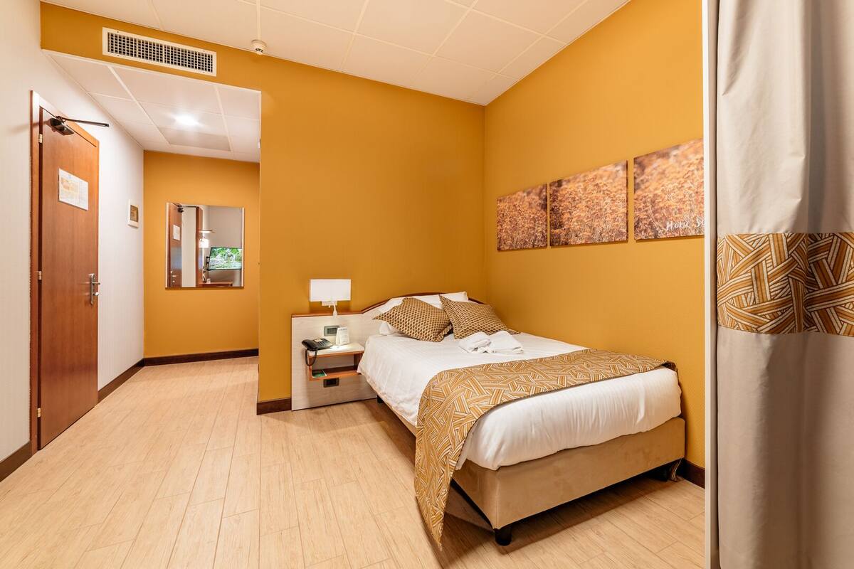 Una habitación de hotel con cama, mesita de noche, lámpara y una obra de arte en la pared.