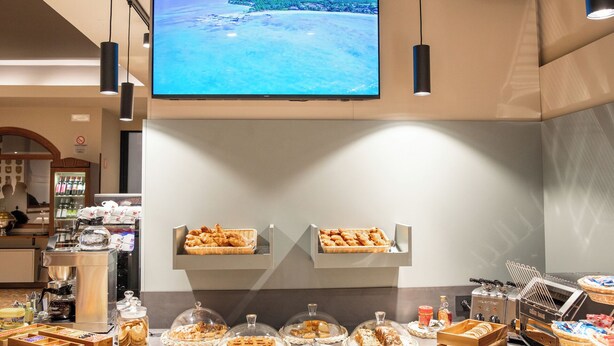 A buffet area with a television displaying a scenic view, various food items, and a coffee station. - Hotel All'Orso (Roncade)