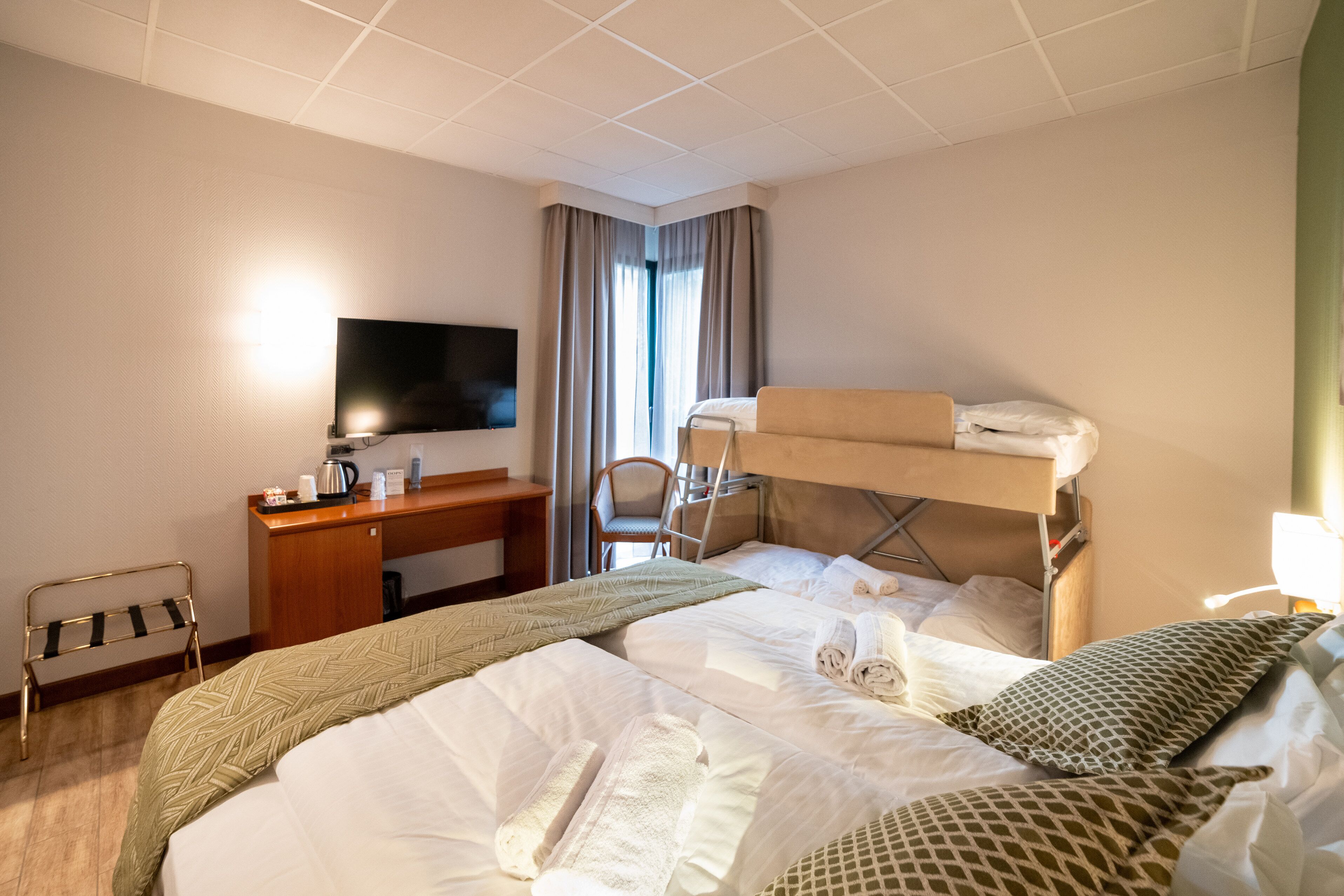 A hotel room with a bunk bed, a desk with a chair, a television, and a window with curtains.