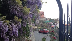 A balcony with a view of a street, a red car, and purple flowers. - Hotel Villa Greta (Taormina)