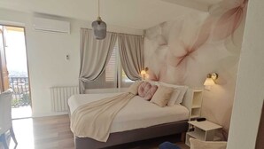 A modern bedroom with a large bed, a nightstand, a chair, and a view of the outdoors. - Hotel Villa Greta (Taormina)