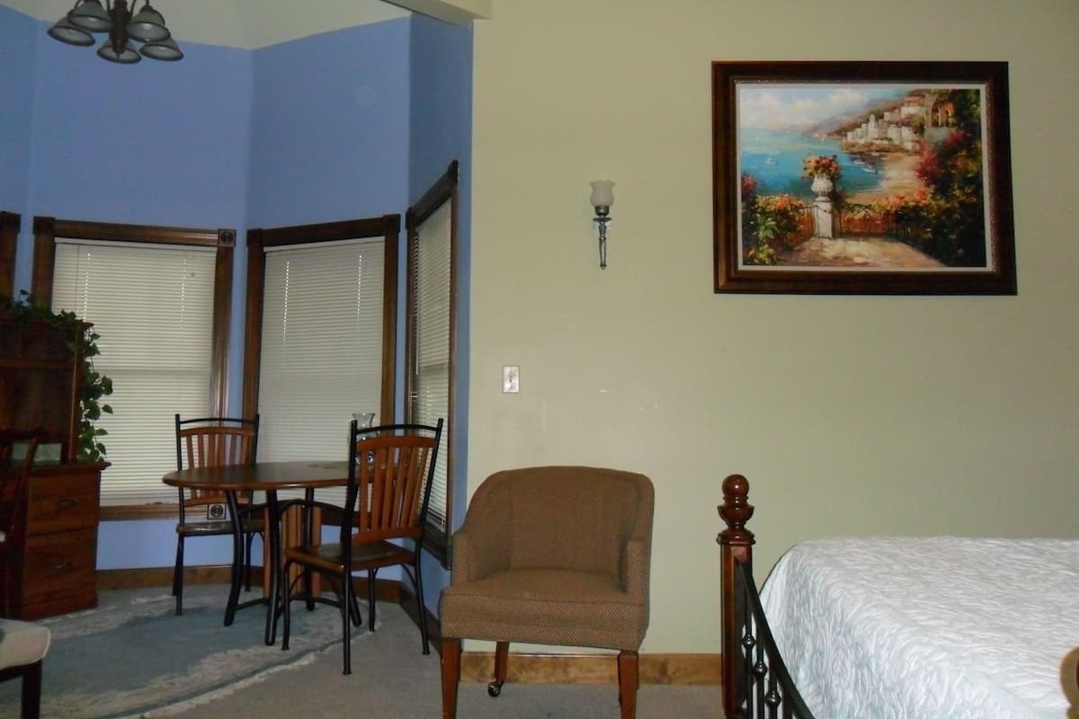 Un dormitorio con cama, una silla, una mesa y un cuadro en la pared.