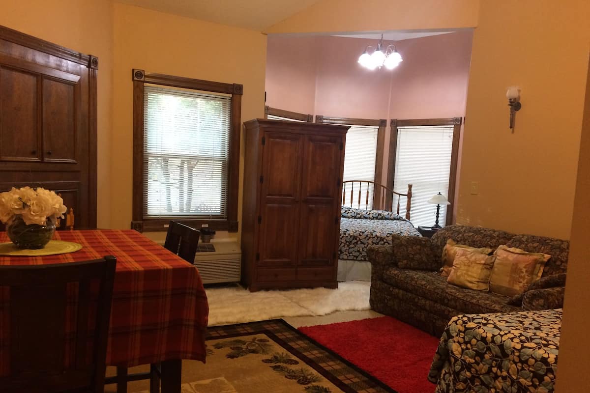 Un comedor con mantel de cuadros rojos, un jarrón con flores y una mesa de madera. En el suelo hay una alfombra estampada, un sofá marrón con una manta floral y una alfombra roja que lleva a un dormitorio con cama y ventana.