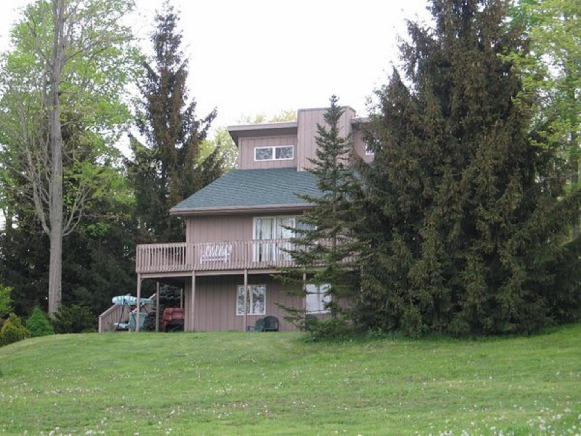 Une maison à deux étages avec un balcon, entourée d’arbres et d’une cour gazonnée.