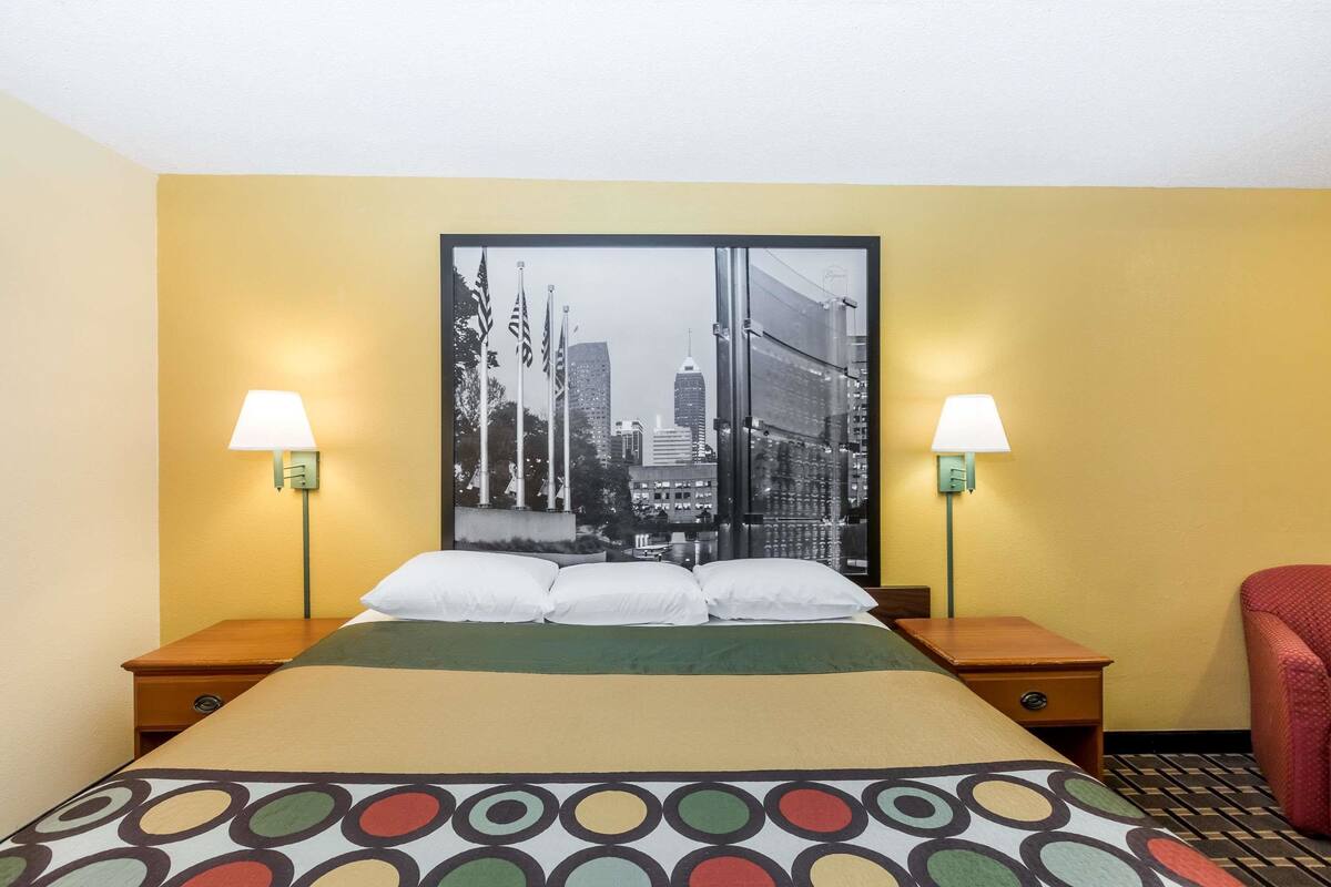 A hotel room with a bed, two bedside lamps, a cityscape poster on the wall, and a red chair.