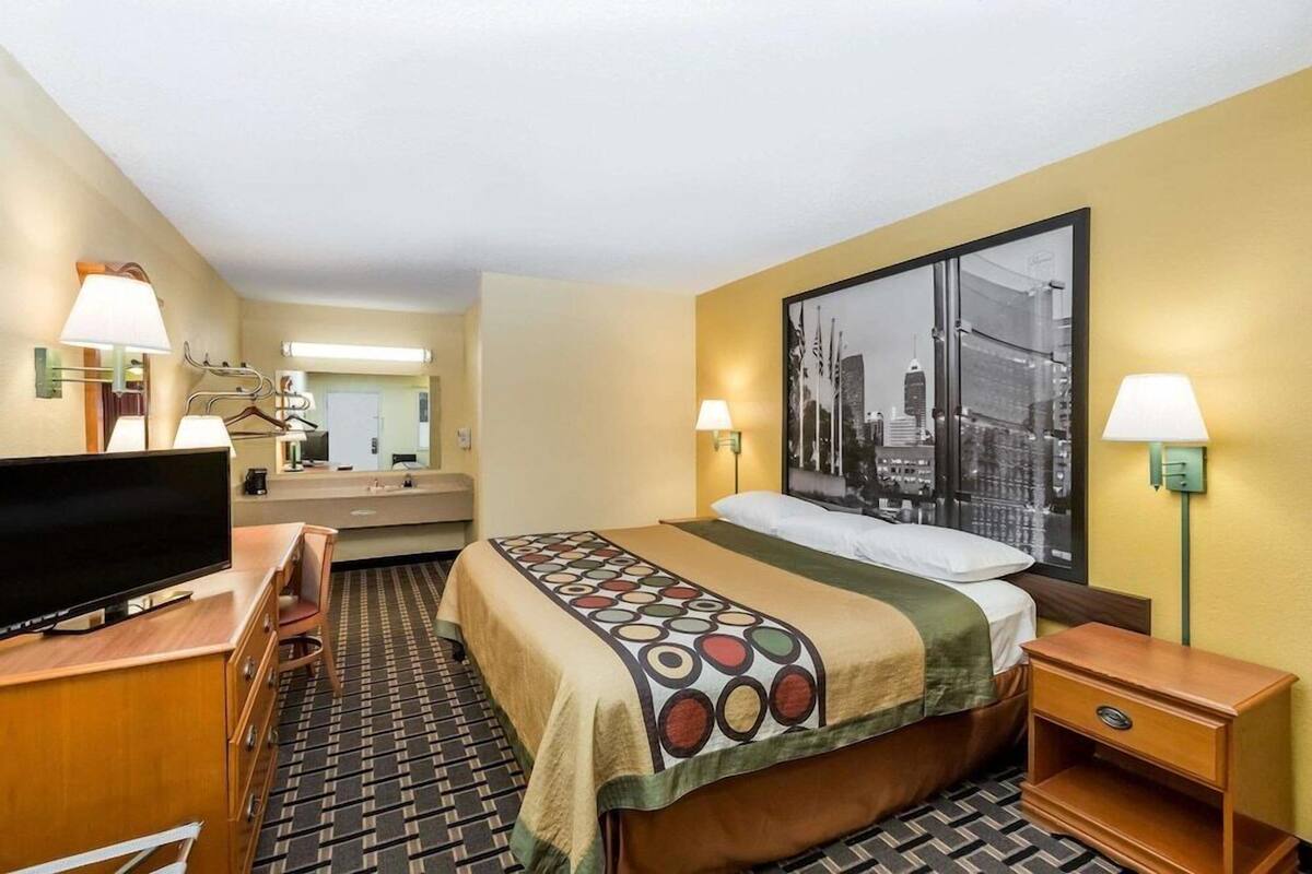 A hotel room with a bed, a television, a desk, a chair, a mirror, and a cityscape painting on the wall.