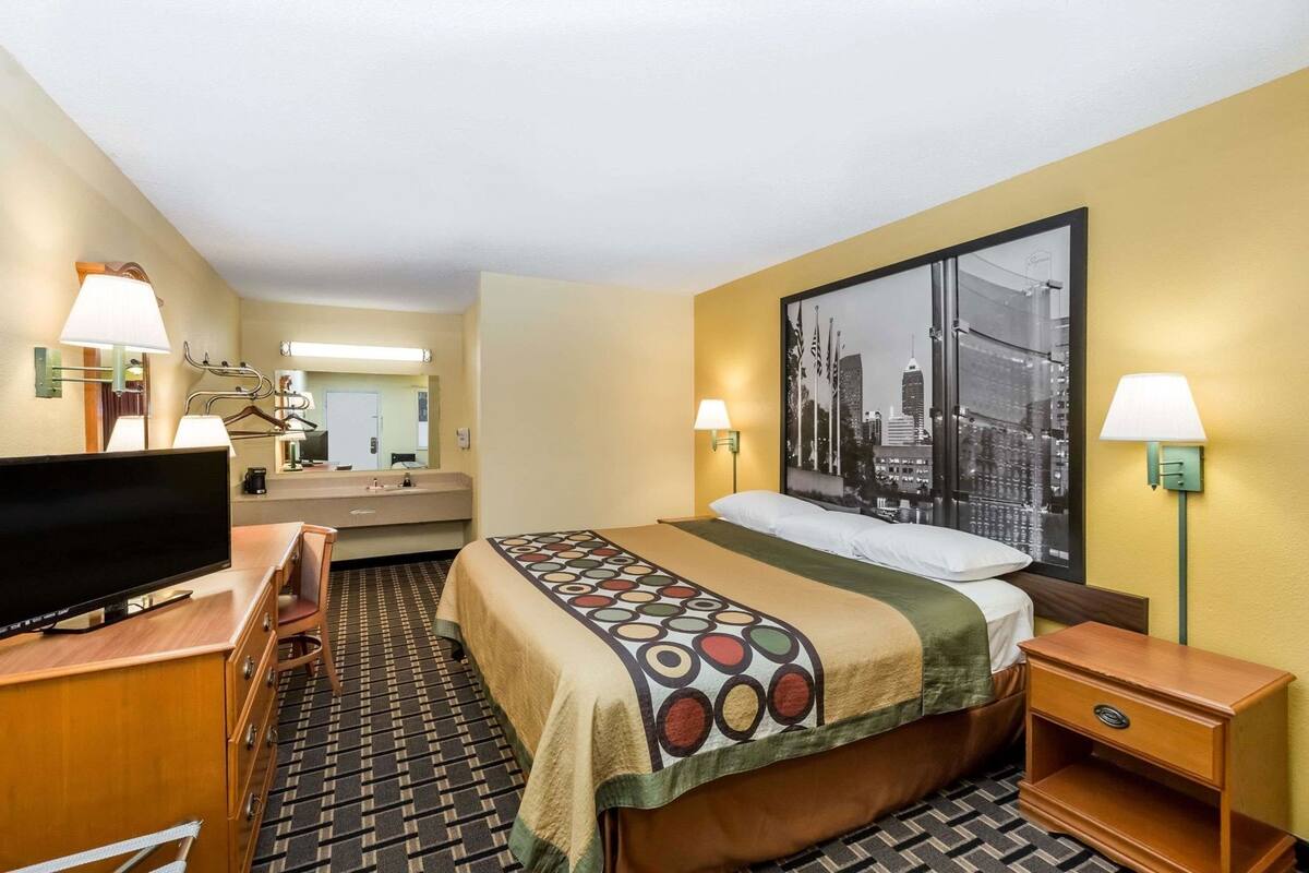 A hotel room with a bed, a TV, a desk, a chair, a dresser, and a large window with a cityscape view.